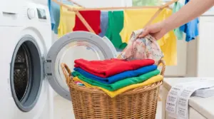 Cestino di panni colorati piegati accanto a una lavatrice aperta con indumenti stesi sullo sfondo